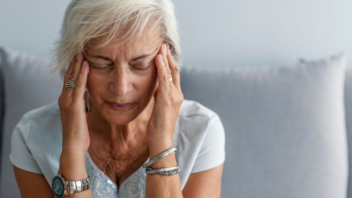 Frau mit grauen Haaren sitzt auf einem Sofa und hält sich mit beiden Händen die Schläfen.
