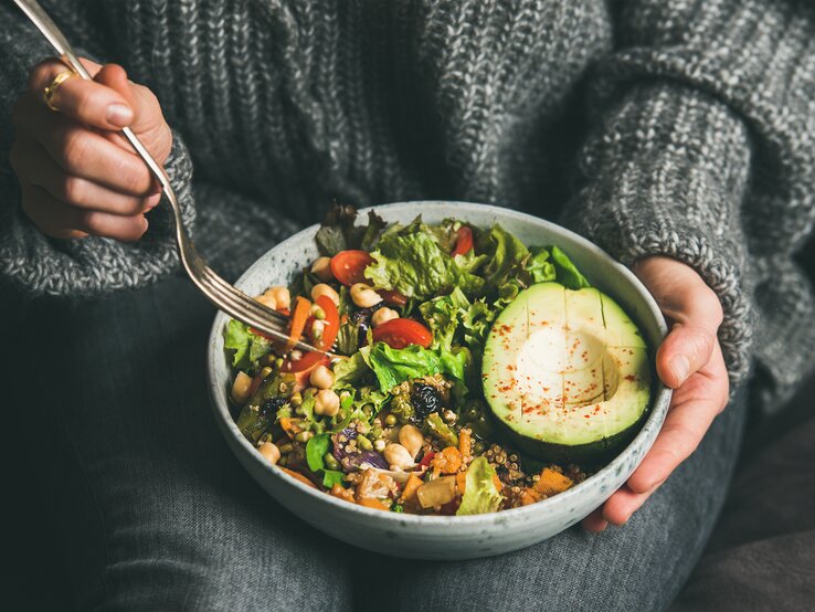 Das Bild zeigt eine Nahaufnahme einer Person, die eine Schale mit einem bunten und gesunden veganen Salat hält. Die Person trägt einen gemütlichen grauen Strickpullover und ist in eine entspannte Haltung versunken, während sie mit einer Gabel in der rechten Hand zum Essen ansetzt. Der Salat besteht aus einer Vielzahl von Zutaten, darunter grüne Blätter, Kichererbsen, Tomaten, Avocado und Körner, die alle frisch und ansprechend aussehen. Die Avocado im Vordergrund ist reif und aufgeschnitten, mit einem Spritzer Gewürz oben drauf. Der Fokus auf den Salat und die Handlungen der Person spiegelt die Wichtigkeit einer achtsamen und gesundheitsbewussten Ernährung wider.
