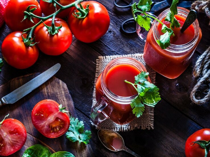 Ein Tisch aus Holz mit einem Brettchen, auf dem ein Messer liegt. Tomaten liegen auf dem Tisch und ein Glas mit Tomatensaft ist von Oben zu sehen. 