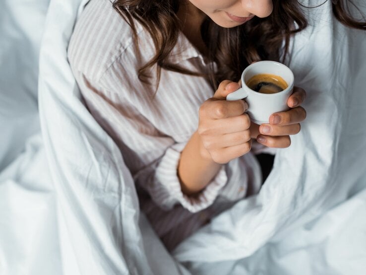Momentaufnahme einer Frau mit dunklem Haar, die morgens gemütlich eine Tasse Espresso im Bett trinkt.