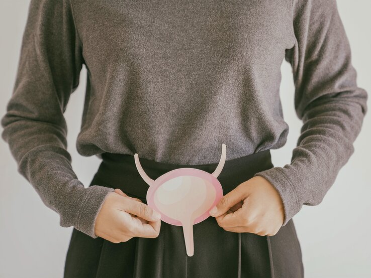 Eine Frau in einem grauen Pullover hält ein rosafarbenes, abstrahiertes Modell einer Blase vor ihrem Bauch.