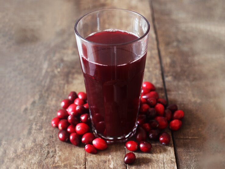 Ein Glas dunkler Cranberrysaft steht auf rustikalem Holztisch, umgeben von frischen roten Cranberries.