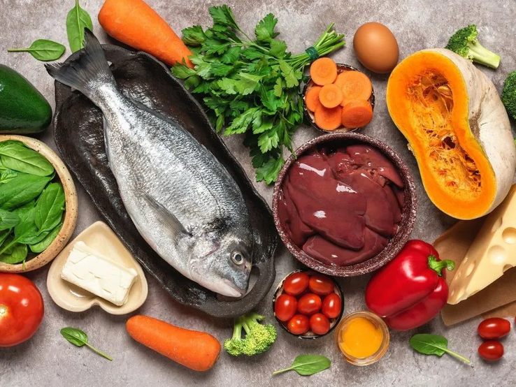 Ein Stillleben mit einer Auswahl an Lebensmitteln, die reich an Nährstoffen sind. Zu sehen sind ein ganzer Fisch auf einem dunklen Teller, eine Schale mit Leberstücken, frisches Gemüse wie Karotten, Paprika, Brokkoli, Spinat, eine Avocado und eine halbierte Kürbishälfte. Außerdem gibt es kleine Tomaten, frische Petersilie, ein Ei, ein Stück Butter und ein Stück Käse. Alles liegt auf einem grauen Untergrund arrangiert.