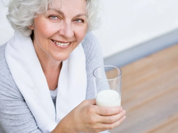 Ältere Frau mit grauen Haaren lächelt und hält ein Glas Milch in der Hand – natürliche Quelle für Vitamin B12.