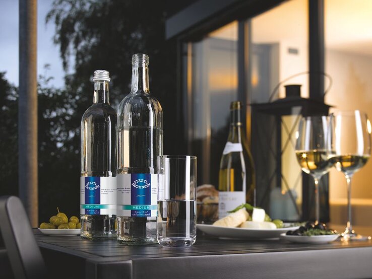 Zwei Flaschen Staatl. Fachingen Medium Mineralwasser auf einem Tisch mit Wein, Käse, Trauben und gefüllten Weingläsern bei stimmungsvoller Abendatmosphäre.