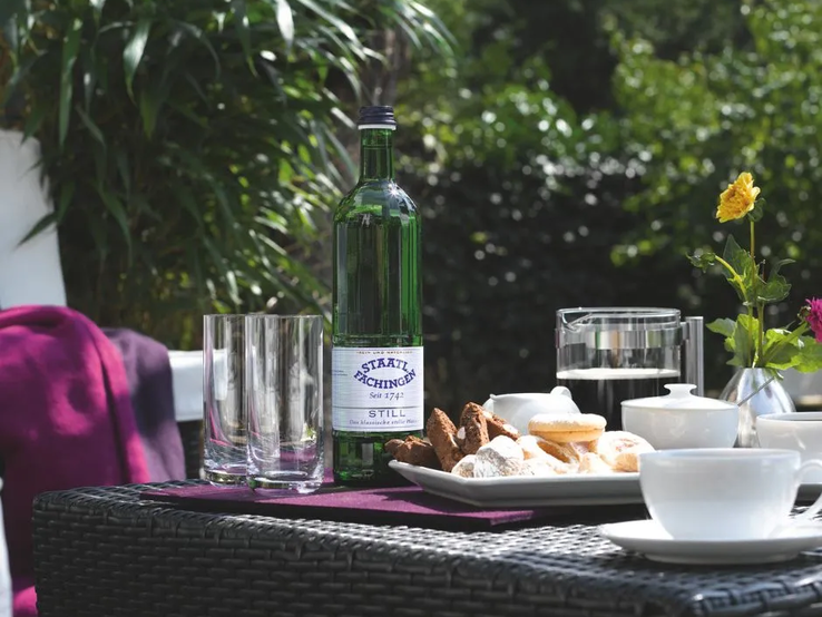Staatl. Fachingen Still Mineralwasser in einer grünen Glasflasche auf einem gedeckten Tisch mit Kaffee, Gebäck und Gläsern im Garten. | © Fachingen Heil- und Mineralbrunnen GmbH