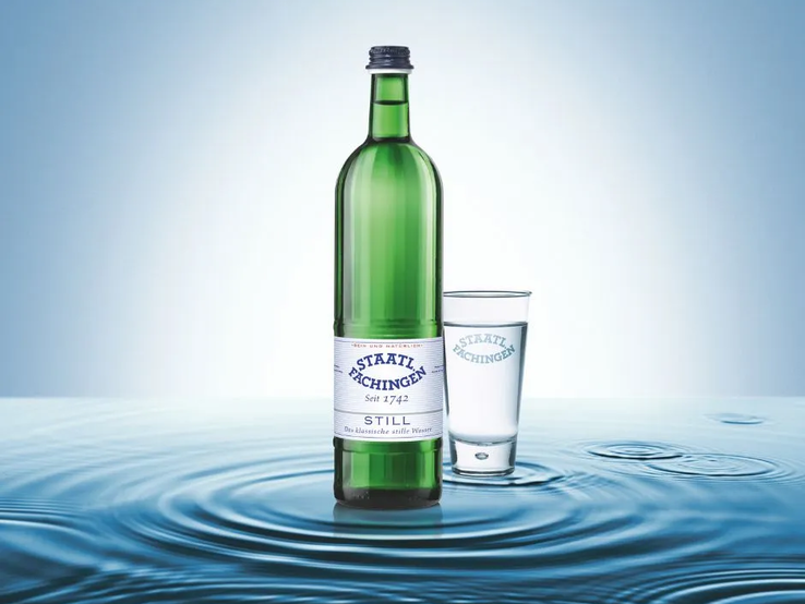 Grüne Glasflasche Staatl. Fachingen Still neben einem Wasserglas, beide stehen auf einer Wasseroberfläche mit kreisförmigen Wellen, vor hellem blauen Hintergrund. | © Fachingen Heil- und Mineralbrunnen GmbH