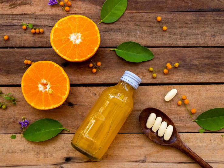 Auf einem Holztisch liegen halbierte Orangen, ein Fläschchen mit Orangensaft und ein Löffel mit Vitamin-C-Tabletten – Symbolbild für hochdosiertes Vitamin C als Nahrungsergänzung im Vergleich zur natürlichen Vitamin-C-Quelle aus Obst.