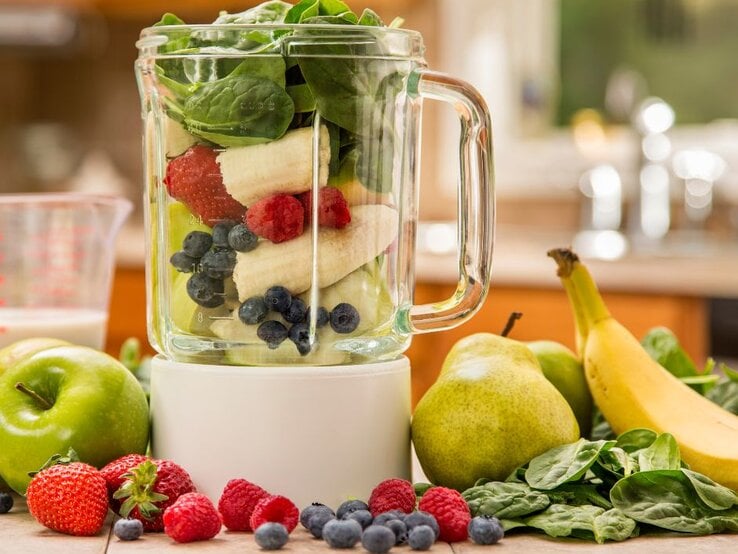 Mixer mit frischem Obst und Gemüse: Bananen, Beeren, Spinat, Äpfel und Birnen liegen auf der Arbeitsplatte und im Glasbehälter – Zutaten für einen gesunden Smoothie.