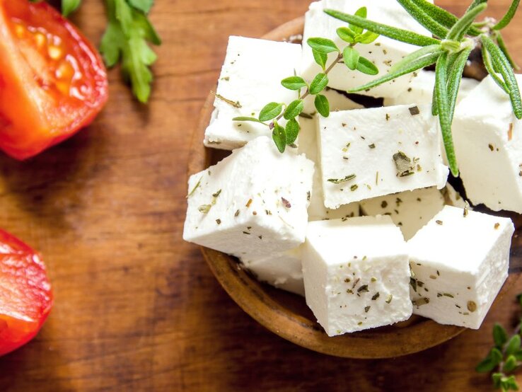 Weiße Feta­würfel mit Kräutern in einer Holzschale, garniert mit frischem Rosmarin und Thymian; daneben liegen halbierte Tomaten und Petersilienblätter auf einem rustikalen Holztisch.