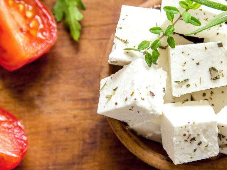 Weiße Feta­würfel mit Kräutern in einer Holzschale, garniert mit frischem Rosmarin und Thymian; daneben liegen halbierte Tomaten und Petersilienblätter auf einem rustikalen Holztisch.