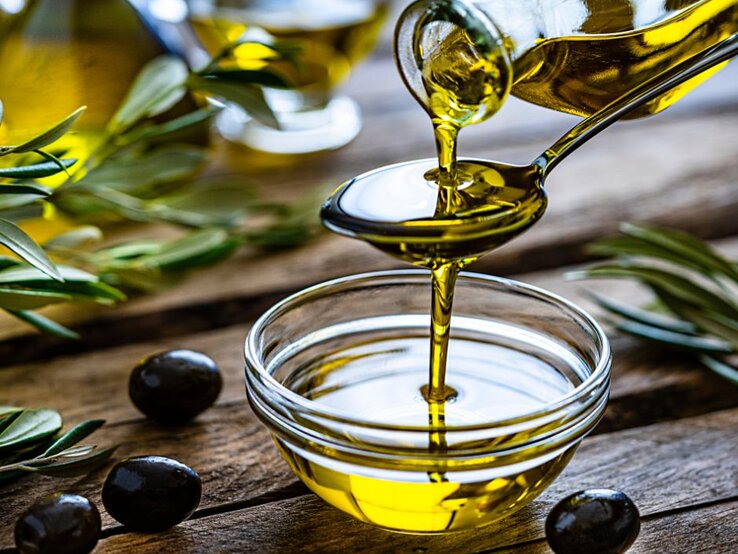 Eine Schale mit goldgelbem Olivenöl steht auf einem Holztisch; darüber gießt jemand frisches Olivenöl aus einer Flasche auf einen Löffel. Rundherum liegen Oliven und Olivenzweige.