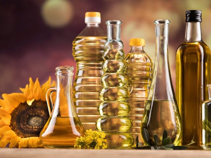 Verschiedene Flaschen mit Speiseöl aus Glas und Kunststoff stehen nebeneinander auf einem Tisch, darunter Sonnenblumenöl und Olivenöl; im Hintergrund sind eine Sonnenblume und warme, unscharfe Farben zu sehen.