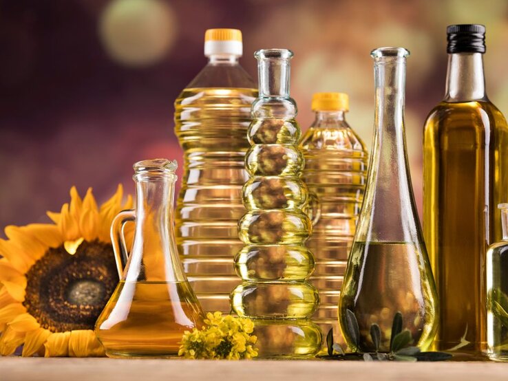 Verschiedene Flaschen mit Speiseöl aus Glas und Kunststoff stehen nebeneinander auf einem Tisch, darunter Sonnenblumenöl und Olivenöl; im Hintergrund sind eine Sonnenblume und warme, unscharfe Farben zu sehen.