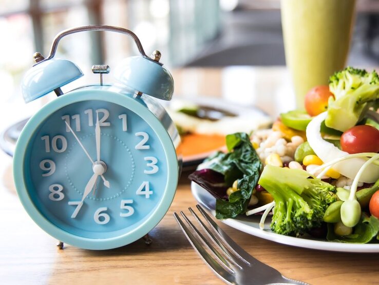 Eine hellblaue Weckeruhr auf einem Holztisch neben einem Teller mit frischem Salat. Der Salat ist bunt und beinhaltet Gemüse wie Brokkoli, Tomaten, Mais und grüne Bohnen. Im Hintergrund ist verschwommen eine weitere Mahlzeit und ein Getränk zu sehen.