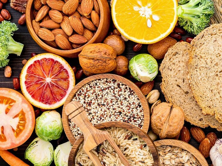 Verschiedene ballaststoffreiche Lebensmittel wie Vollkornbrot, Nüsse, Samen, Reis, Quinoa, Hülsenfrüchte, Brokkoli, Rosenkohl, Karotten und Zitrusfrüchte liegen dekorativ auf einem dunklen Untergrund.