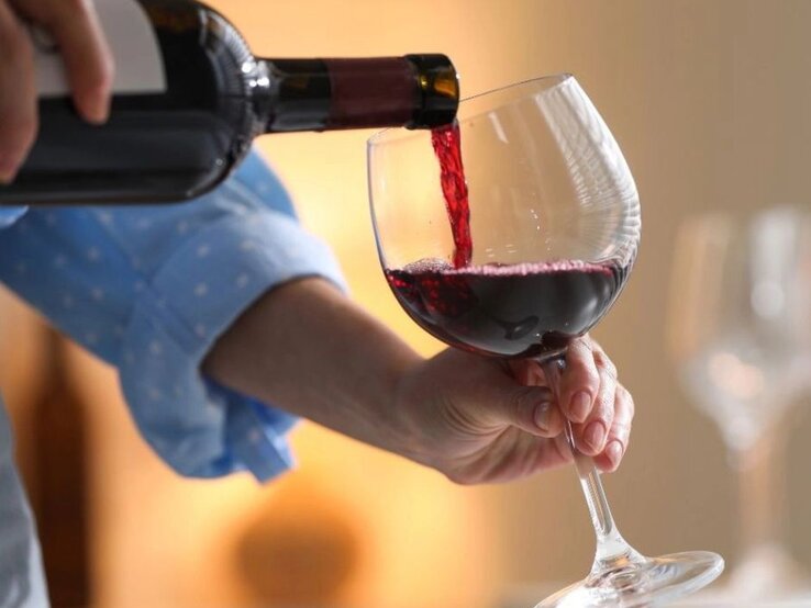 Eine Person gießt roten Wein aus einer Flasche in ein großes Weinglas; die Hand hält das Glas leicht schräg, im Hintergrund ist eine warme, wohnliche Atmosphäre zu sehen.