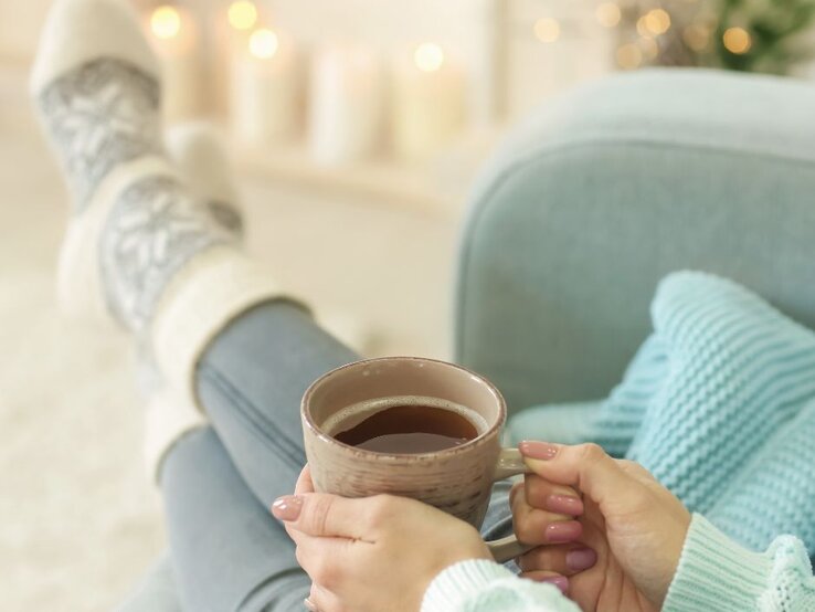 Frau in gemütlicher Kleidung sitzt entspannt auf einem Sofa, trägt dicke Stricksocken und hält eine Tasse Tee oder Kaffee in den Händen, im unscharfen Hintergrund warme Kerzen- und Lichterdekoration.