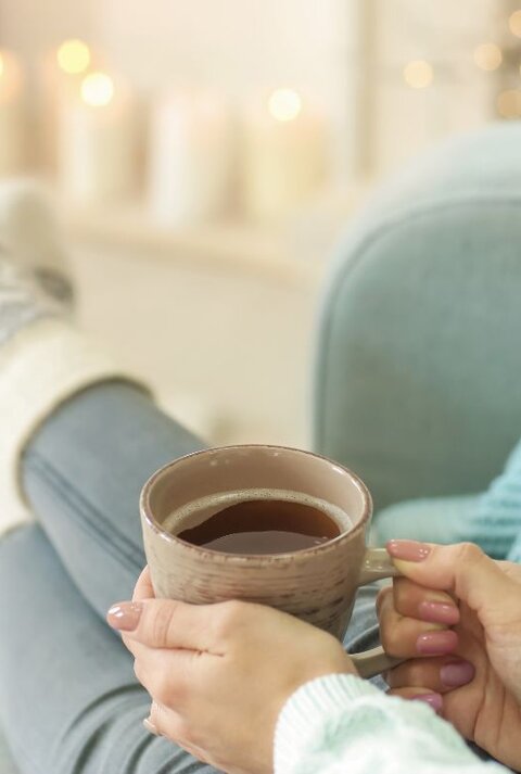 Frau in gemütlicher Kleidung sitzt entspannt auf einem Sofa, trägt dicke Stricksocken und hält eine Tasse Tee oder Kaffee in den Händen, im unscharfen Hintergrund warme Kerzen- und Lichterdekoration.