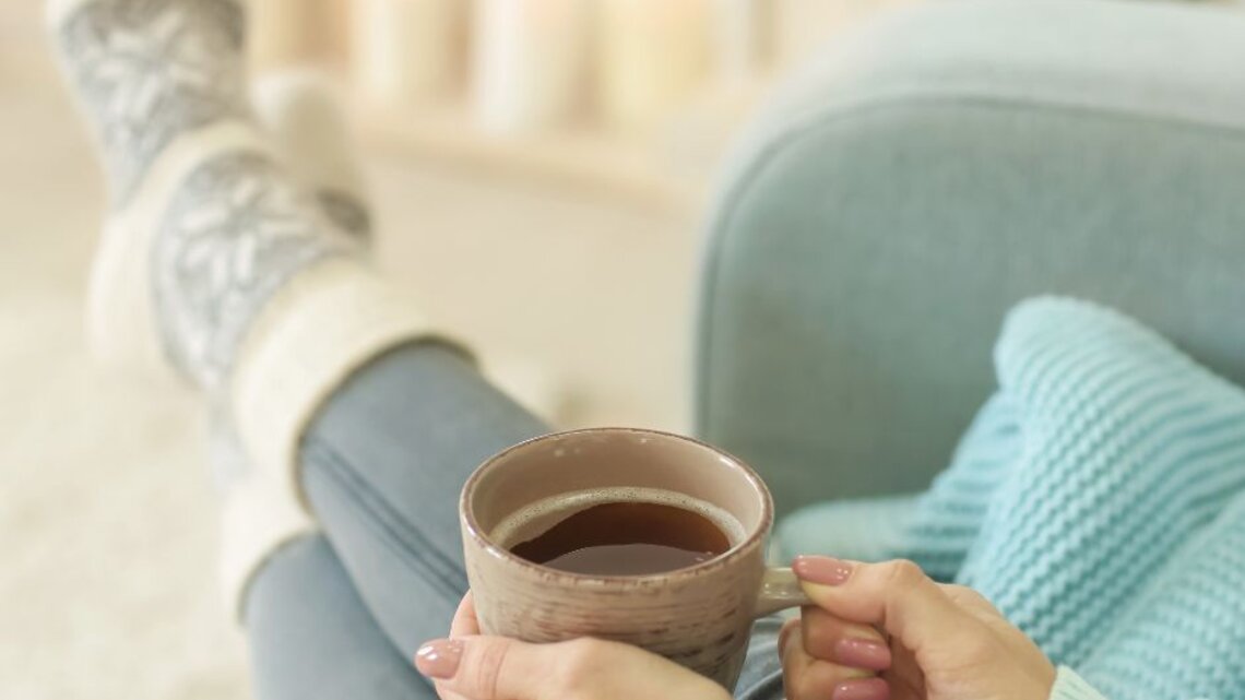 Frau in gemütlicher Kleidung sitzt entspannt auf einem Sofa, trägt dicke Stricksocken und hält eine Tasse Tee oder Kaffee in den Händen, im unscharfen Hintergrund warme Kerzen- und Lichterdekoration.
