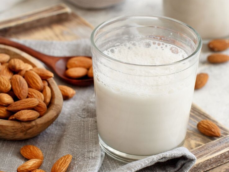 Ein Glas frisch zubereitete Mandelmilch steht auf einem Holzbrett, umgeben von ganzen Mandeln in einer Holzschale sowie einer Stoffserviette. Im Hintergrund ist ein weiteres Glas mit Mandelmilch unscharf zu erkennen.