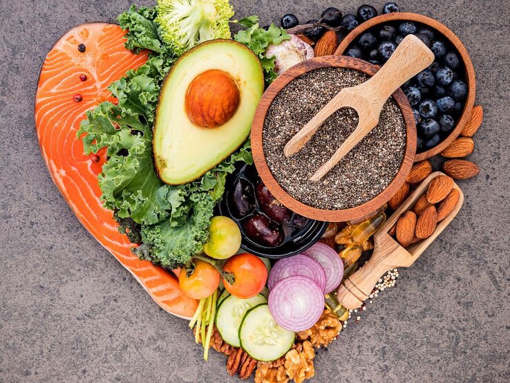 Ein herzförmiges Arrangement aus gesunden Lebensmitteln, darunter Lachs, Avocado, Grünkohl, Chiasamen, Blaubeeren, Mandeln, Walnüsse, Kirschtomaten, rote Zwiebeln, Gurkenscheiben, Brokkoli und Knoblauch. Die Zutaten sind farbenfroh präsentiert und symbolisieren eine ausgewogene Ernährung.   Fragen  Erklären
