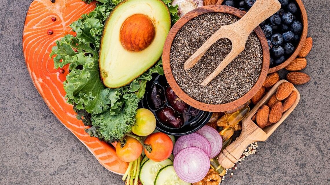 Ein herzförmiges Arrangement aus gesunden Lebensmitteln, darunter Lachs, Avocado, Grünkohl, Chiasamen, Blaubeeren, Mandeln, Walnüsse, Kirschtomaten, rote Zwiebeln, Gurkenscheiben, Brokkoli und Knoblauch. Die Zutaten sind farbenfroh präsentiert und symbolisieren eine ausgewogene Ernährung.   Fragen  Erklären