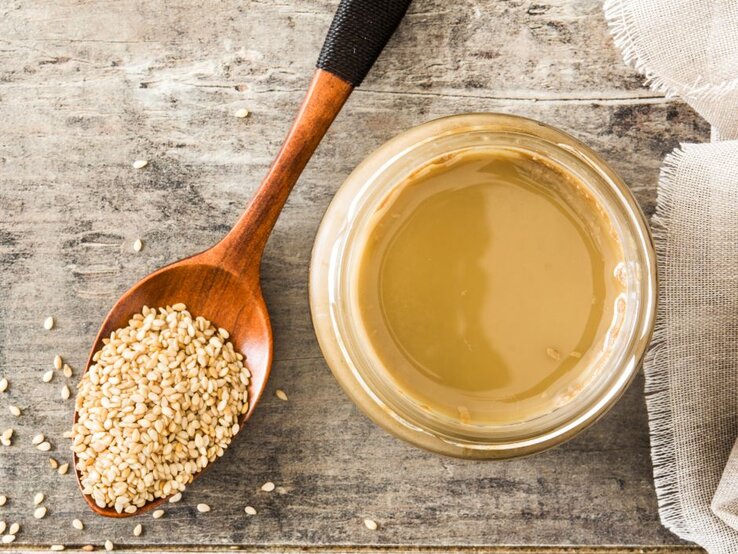 Holzlöffel mit Sesamsamen neben einem Glas Tahini auf Tisch. | © estefaniavizciano via Canva.com