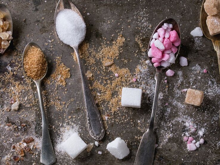 Mehrere Löffel mit unterschiedlicher Zuckerart – weißer Zucker, brauner Zucker und pink-weiße Zuckerstückchen – liegen auf einer dunklen Oberfläche, umgeben von verstreuten Zuckerwürfeln und -krümeln.