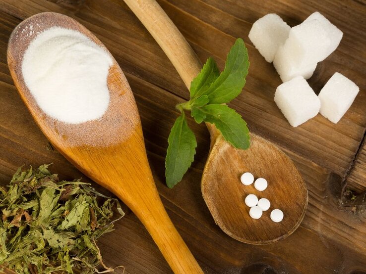 Holzlöffel mit Suüßstoffen auf einer braunen Tischplatte. Daneben Zuckerwürfel. 