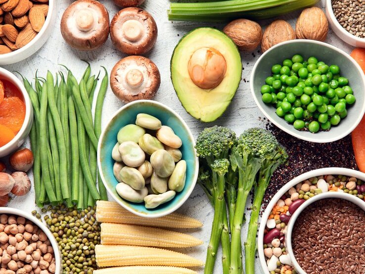 Von oben fotografierte Vielfalt pflanzlicher Lebensmittel—Erbsen, Bohnen, Linsen, Avocado, Brokkoli, Pilze, grüne Bohnen, Nüsse, Samen und Babymais—auf hellem Untergrund.