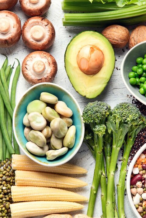 Von oben fotografierte Vielfalt pflanzlicher Lebensmittel—Erbsen, Bohnen, Linsen, Avocado, Brokkoli, Pilze, grüne Bohnen, Nüsse, Samen und Babymais—auf hellem Untergrund.