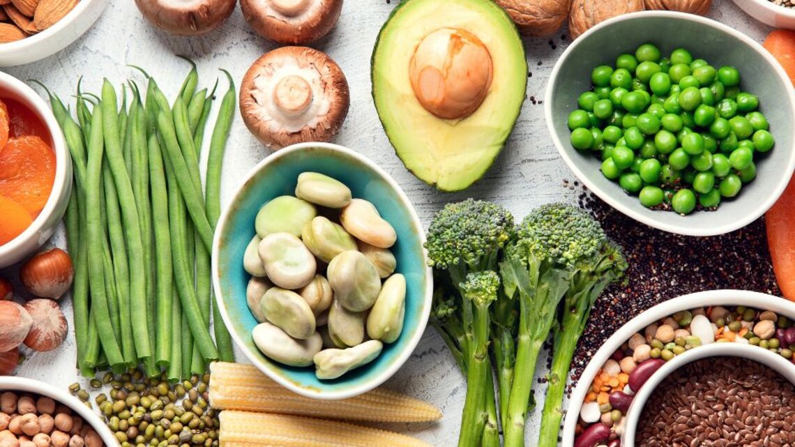 Von oben fotografierte Vielfalt pflanzlicher Lebensmittel—Erbsen, Bohnen, Linsen, Avocado, Brokkoli, Pilze, grüne Bohnen, Nüsse, Samen und Babymais—auf hellem Untergrund.