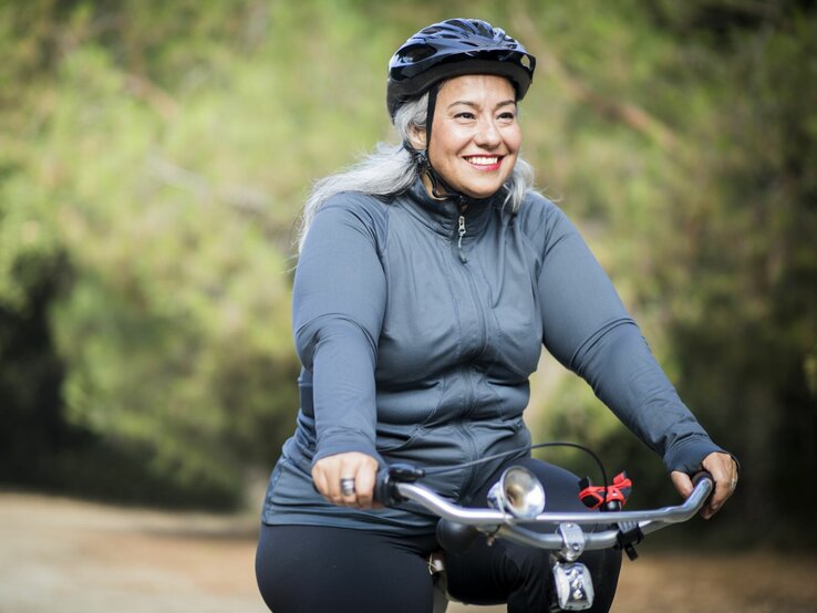 Eine fröhliche ältere Frau mit grau melierten Haaren fährt Rad in einem Freizeitoutfit. Sie trägt einen Fahrradhelm und ein langärmeliges Oberteil und lächelt, während sie auf einem Weg umgeben von grüner Vegetation radelt