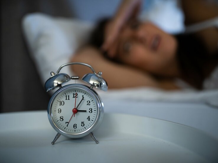 Das Foto zeigt einen klassischen Wecker im Vordergrund, der auf einem Nachttisch steht, mit einer unscharfen Person im Hintergrund, die im Bett liegt und sich anscheinend mit Schlafschwierigkeiten herumschlägt. Die Person hält sich eine Hand an die Stirn in einer Geste von Frustration oder Erschöpfung. Der Fokus auf den Wecker unterstreicht das Thema Schlaflosigkeit und die damit verbundenen Herausforderungen.