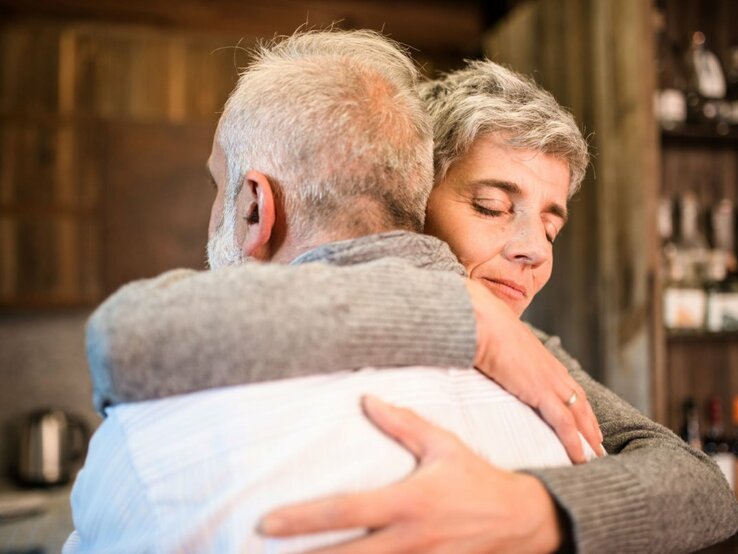 Das Foto zeigt zwei ältere Personen, die sich in einer herzlichen Umarmung befinden. Die Frau, die in die Kamera blickt, hat kurzes graues Haar und ein geschlossenes, friedliches Lächeln, während der Mann ihr seine Zuneigung zeigt, indem er sie fest hält. Das Paar scheint sich in einer häuslichen oder gemütlichen Umgebung zu befinden, was durch unscharfe Hintergrundelemente wie Regale mit verschiedenen Gegenständen angedeutet wird. Das Bild strahlt Wärme, Liebe und Fürsorge aus.