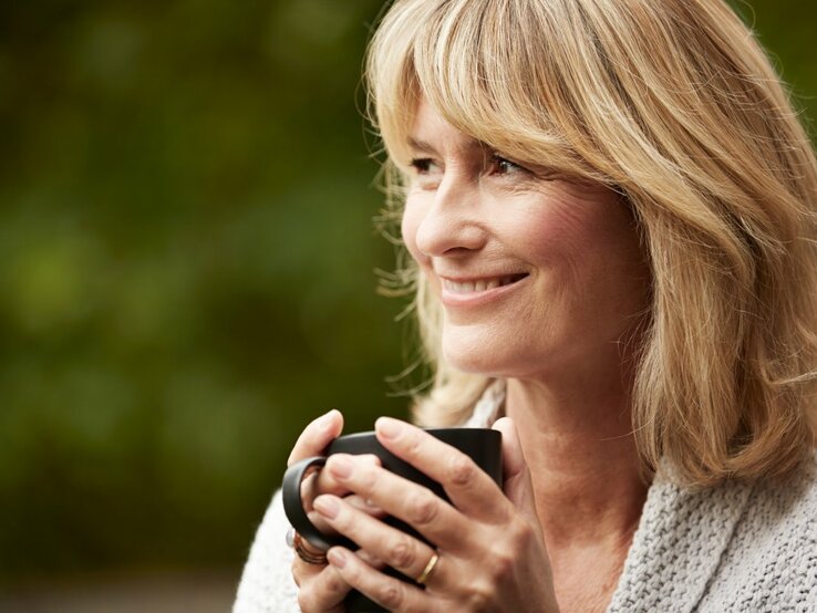 Das Foto zeigt eine reife Frau mit blonden Haaren, die lächelnd in die Ferne schaut, während sie eine dunkle Teetasse in ihren Händen hält. Sie trägt einen gemütlichen grauen Strickpullover und scheint sich in einem entspannten Moment im Freien zu befinden, mit einem unscharfen grünen Hintergrund, der auf einen Garten oder Park hindeuten könnte. Die sanfte Mimik der Frau strahlt Ruhe und Zufriedenheit aus.