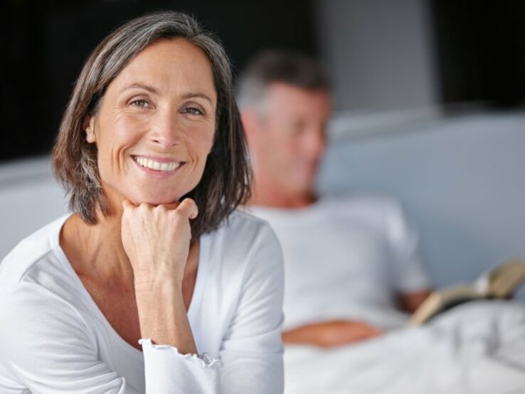Reife Frau sitzt auf Bett und schaut in die Kamera während ihr Mann im Hintergrund liest | ©  iStock/laflor