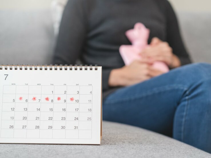 Das Bild zeigt eine Person, die auf einem Sofa sitzt und eine Wärmflasche an den Unterleib drückt, ein Zeichen für Menstruationsbeschwerden. Im Vordergrund steht ein Kalender auf einem Tisch, mit einigen Tagen markiert, was darauf hindeuten könnte, dass die Person ihre Menstruationszyklen verfolgt. Der Kalender fokussiert auf den Monat Juli, und die markierten Tage scheinen die erwartete Periode darzustellen.