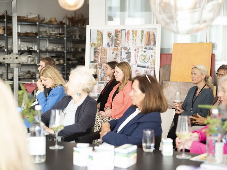Frauen unterschiedlichen Alters sitzen aufmerksam in heller Küche, einige mit Getränken, im Hintergrund Regale und Food-Collagen. | © BILD der FRAU