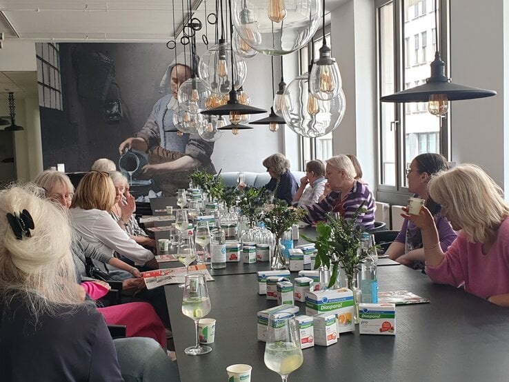 Eine Gruppe älterer Frauen sitzt tagsüber an langem Tisch mit Diasporal-Produkten, Zitronenwasser und Blumen unter Glaskugellampen. | © BILD der FRAU