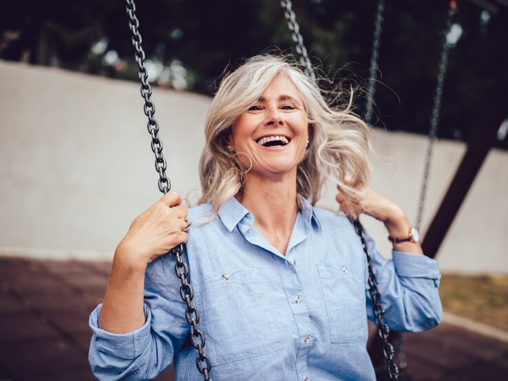 In der Postmenopause verschwinden die schlimmsten Beschwerden der Wechseljahre und ein neuer Lebensabschnitt beginnt. Frau sitzt auf einer Schaukel und lacht.