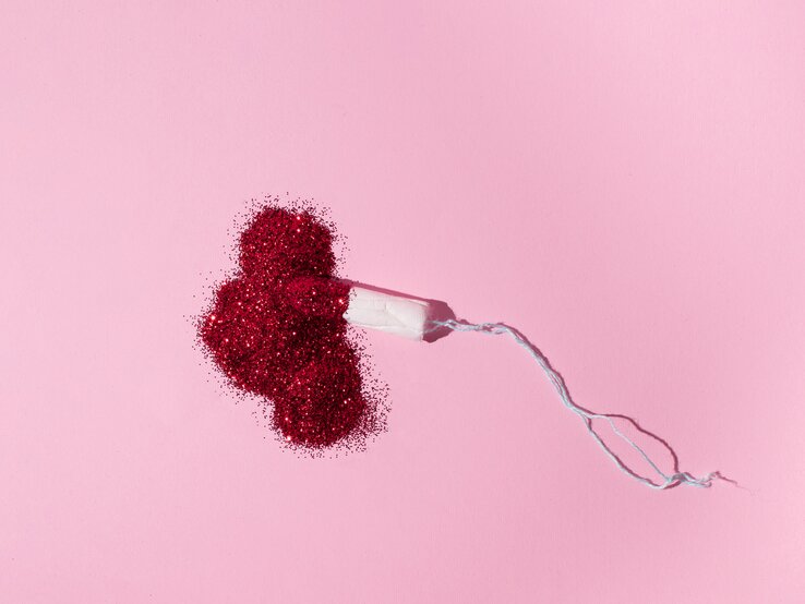 Blutungen in den Wechseljahren kommen oft ungeplant und überraschend. Tampon liegt auf rosa Untergrund in einem Haufen aus rotem Glitzer.