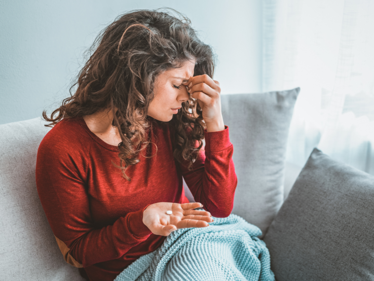 Frau mit Schmerzen auf dem Sofa und Pille in der Hand Junge Frau mit langen braunen Locken sitzt erschöpft auf dem Sofa, hält Tabletten in der Hand und reibt sich die Stirn.