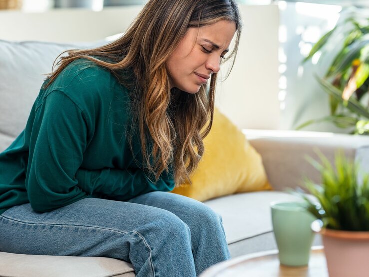 Eine Frau, die einen dunkelgrünen Pullover trägt und braune Haare hat, sitzt auf einem hellen Sofa und hält sich mit schmerzverzerrtem Gesicht den linken Bauchbereich. Sie scheint starke Schmerzen zu haben.