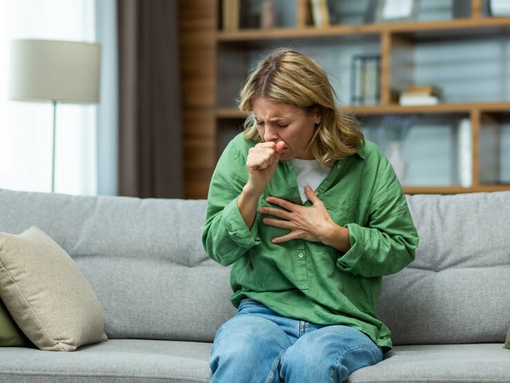 Das Bild zeigt eine Frau mittleren Alters, die auf einem Sofa sitzt und sich mit einer Hand auf die Brust fasst, während sie mit der anderen Hand ihren Mund bedeckt, als würde sie husten. Sie wirkt besorgt oder unwohl, möglicherweise aufgrund des Reizhustens oder verschleimten Hustens. Die Umgebung deutet auf ein häusliches Wohnzimmer hin, mit einem modernen, ruhigen Hintergrund, der das Hauptaugenmerk auf die Frau lenkt. Ihre Kleidung ist leger, mit einem grünen Hemd und blauen Jeans, was darauf hindeutet, dass sie sich in einer entspannten Umgebung befindet.