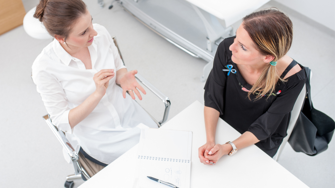 Ärztin im Gespräch mit einer Patientin, beide sitzen an einem Tisch, Perspektive von oben.