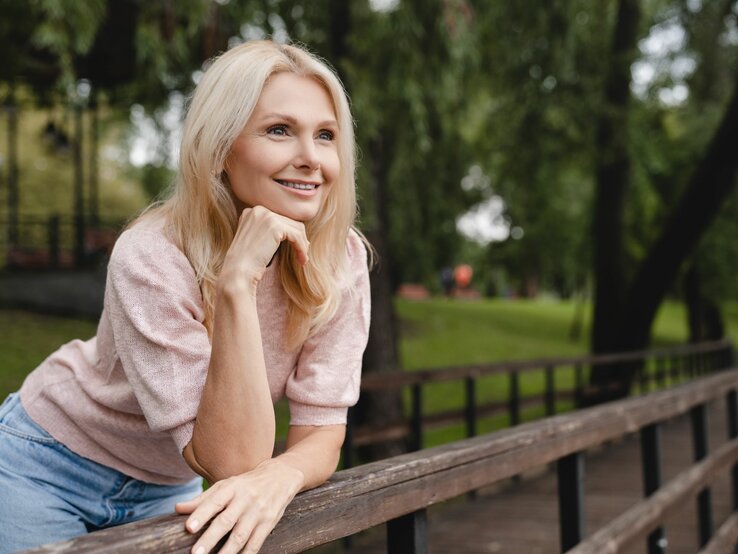 Das Foto zeigt eine lächelnde blonde Frau, die entspannt im Freien sitzt und sich an eine hölzerne Brüstung lehnt. Sie trägt ein rosa Oberteil und eine blaue Jeans. Ihr Blick ist nachdenklich und sie scheint etwas in der Ferne zu betrachten. Der Hintergrund ist unscharf, mit Bäumen und Grünflächen, was auf einen Park oder eine ähnliche natürliche Umgebung hindeutet. Die entspannte Pose und das freundliche Lächeln der Frau vermitteln eine ruhige und zufriedene Ausstrahlung.