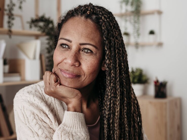 Das Foto zeigt eine Frau mit einem nachdenklichen Gesichtsausdruck, die ihre Wange mit der Hand stützt. Sie hat lange, geflochtene Haare und trägt einen gemütlichen Strickpullover. Im Hintergrund ist ein helles und modern eingerichtetes Zimmer mit Pflanzen und Regalen zu erkennen. Ihr Blick scheint in die Ferne gerichtet, was darauf hindeuten könnte, dass sie in Gedanken versunken ist.