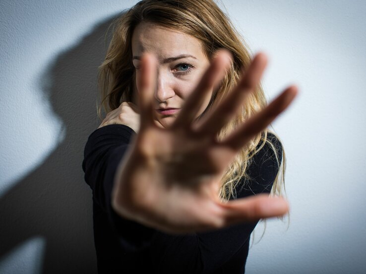 Das Foto zeigt eine junge Frau, die ihre Hand zur Kamera hin ausstreckt, als ob sie versucht, die Aufnahme zu blockieren oder sich zu schützen. Ihr Gesichtsausdruck wirkt besorgt oder ängstlich. Die Beleuchtung erzeugt einen dramatischen Kontrast und Schatten an der Wand hinter ihr, was die emotionale Intensität des Bildes verstärkt. Es könnte eine Darstellung von Abwehr, Privatsphäre oder einer emotionalen Grenze sein.